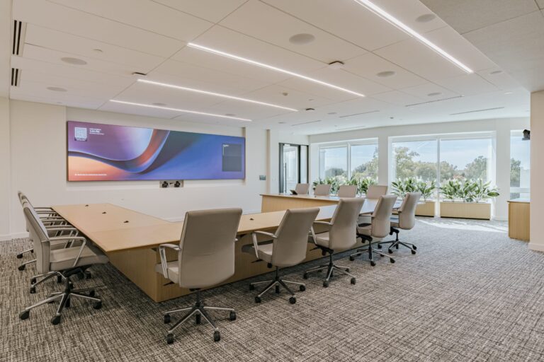 A modern conference room featuring a large rectangular wooden table surrounded by several ergonomic chairs on wheels. There is a large digital display on the wall, and the room is well-lit with natural light coming from large windows adorned with potted plants. The ceiling has linear LED lighting, and the floor is carpeted in a gray pattern.