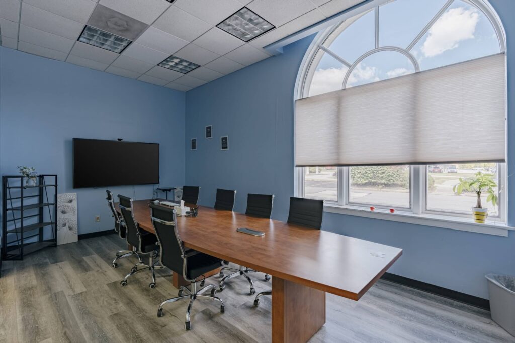 A modern conference room with blue walls, featuring a large wooden table surrounded by black office chairs. The room has a large arched window with vertical blinds partially closed, allowing light to filter in. A flat screen monitor is mounted on one wall, and a potted plant is placed on the windowsill. The ceiling has recessed lighting, and the floor has a wood-style finish.