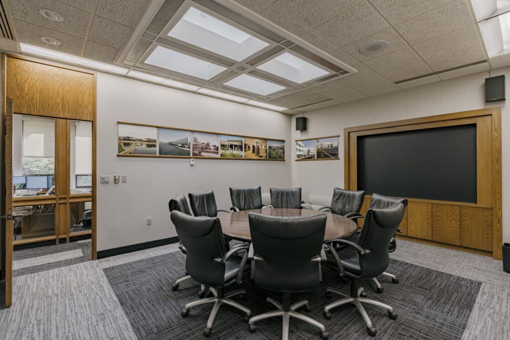 A modern conference room featuring a round wooden table surrounded by seven black leather swivel chairs on a carpeted floor. The walls are adorned with framed landscape photographs, and there is a large blank screen set in a wooden frame on one wall. On the left, a wooden door, partially open, reveals an adjoining office space.