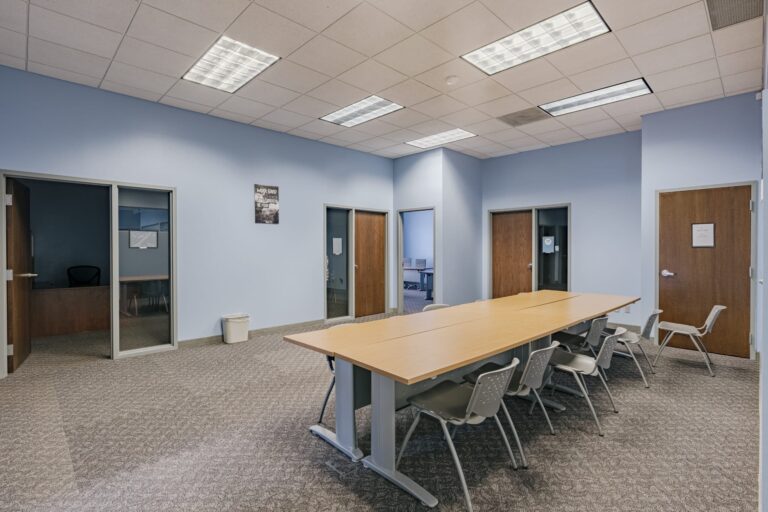 A spacious, modern conference room featuring a long wooden table surrounded by several grey chairs. The room has blue walls and a carpeted floor with ceiling lights providing bright illumination. There are multiple wooden doors and a glass-paneled office visible in the background.