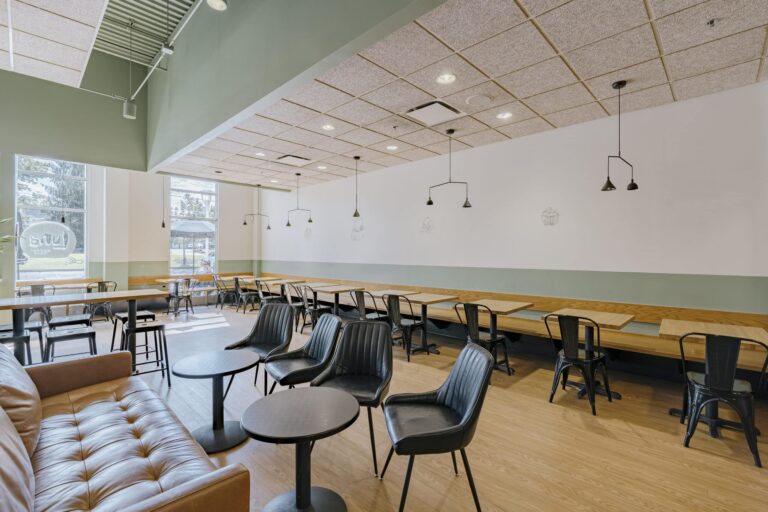 A modern cafe interior featuring a mix of seating options, including leather couches, round black tables with black chairs, and long communal wooden tables with several black metal chairs. The space has large windows with natural light streaming in, green and white walls, stylish ceiling lights, and a contemporary aesthetic.