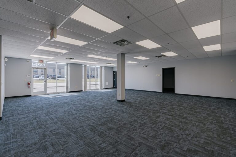 A spacious and modern empty office space with grey carpeting, white ceiling tiles, and fluorescent lighting. Large glass windows and an exit door are visible, allowing natural light to enter. There is a fire extinguisher mounted on the wall next to the door, and an open doorway leading to another room.