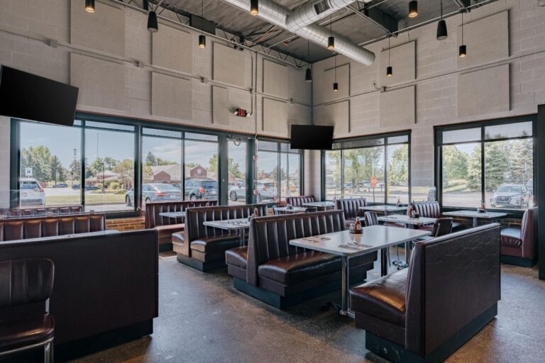 Interior of a modern restaurant featuring empty booth seating with brown leather upholstery. Large glass windows provide a view of the parking area and trees outside. Tables are set with utensils and condiments, and the ceiling features exposed ductwork with hanging lights.