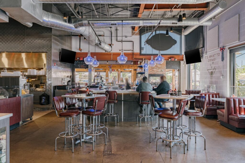 Interior of a modern restaurant with an industrial design featuring exposed ducts and pipes on the ceiling. The space includes several high tables with red leather chairs and a bar area at the center with hanging blue pendant lights. Two patrons are seated at the bar, and a kitchen is visible in the background. Large windows allow natural light to fill the room.