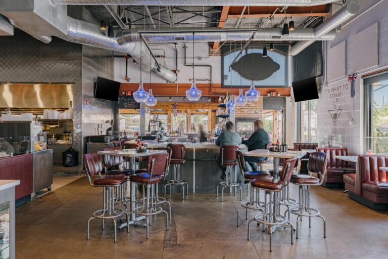 Interior of a modern restaurant with an industrial design featuring exposed ducts and pipes on the ceiling. The space includes several high tables with red leather chairs and a bar area at the center with hanging blue pendant lights. Two patrons are seated at the bar, and a kitchen is visible in the background. Large windows allow natural light to fill the room.