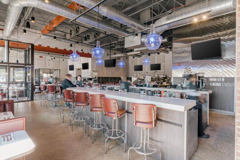 A modern restaurant interior with an industrial design featuring exposed ductwork and large blue pendant lights. The image shows a long bar with red swivel chairs and patrons seated. The counter area is equipped with various bar tools and appliances, and there are several wall-mounted televisions. The atmosphere appears lively with people actively serving and engaging at the bar.