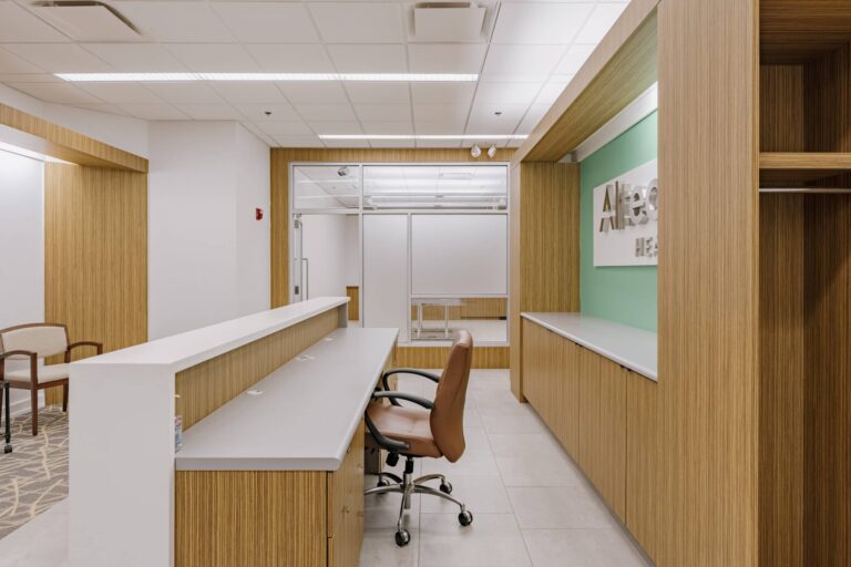 A modern medical office reception area featuring a long white and wood reception desk with a brown office chair. The walls are adorned with light wood paneling and a green accent wall with a partially visible logo. The ceiling has recessed lighting, and a seating area with beige chairs is visible to the left.