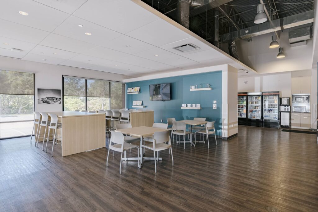 A modern office break room featuring wooden tables and chairs, a teal accent wall with a mounted TV, and large windows providing natural light. The area includes a kitchenette with refrigerated drinks, microwaves, and white cabinetry.