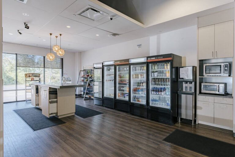 A modern office break room featuring a central island with a countertop and open storage underneath. There are several tall refrigerated display cases stocked with various drinks, including sodas and water bottles. On the right, built-in cabinets house multiple microwaves, and a touchless water dispenser is visible. The room is well-lit with natural light from large windows, and pendant lights hang from the ceiling. The flooring is finished with wood, and door mats are positioned in key areas.