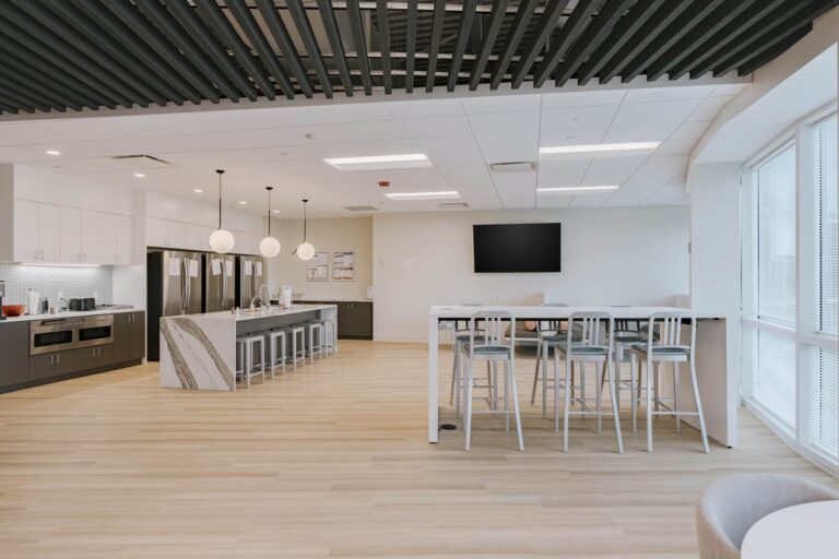 A spacious and modern office breakroom featuring a large kitchen area with white cabinetry, multiple stainless steel refrigerators, and an island with barstools. The room includes a flat-screen TV on the wall, a long table with high chairs, and ample natural light from large windows.