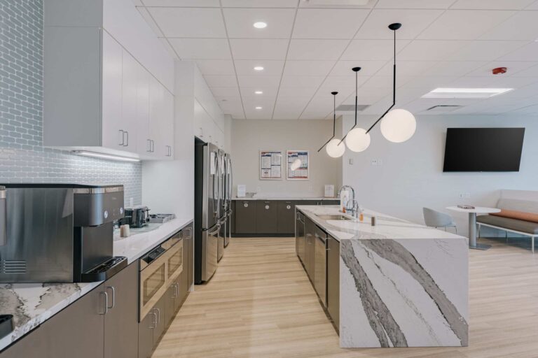 A modern office breakroom featuring sleek cabinetry and stainless steel appliances. The space includes a coffee machine, microwave, and multiple refrigerators. A stylish kitchen island with a marbled countertop is accented by contemporary pendant lighting. In the background, a seating area with a table and TV adds convenience for employees.