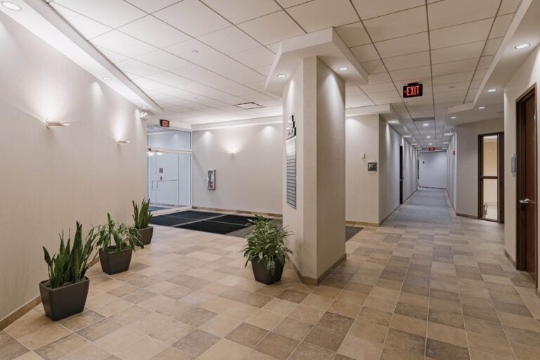 A modern office building hallway with beige tiled floors and white walls. The space is brightly lit with ceiling lights, and there are several potted green plants placed along the walls. On the left side, there is a glass entry door with an exit sign above it, and on the right, a long corridor extends into the distance with a gray carpeted floor.