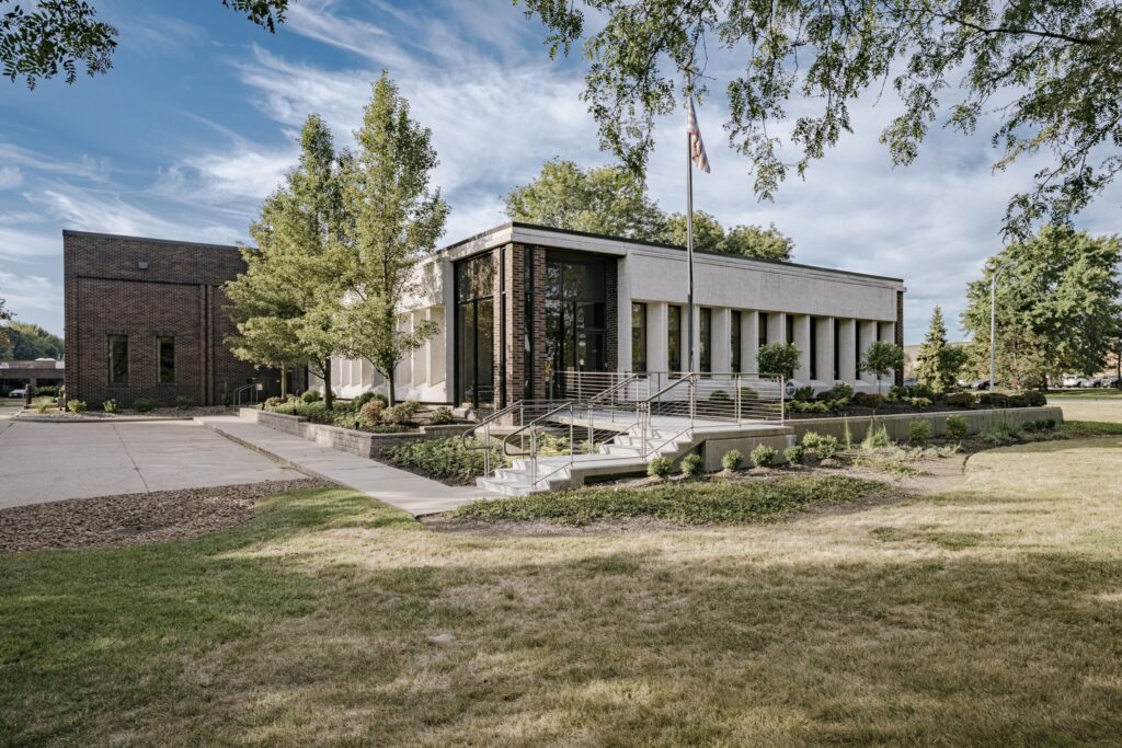 A modern office building with large windows and a mix of brick and concrete exterior surrounded by landscaped grounds with pathways and trees. The American flag is flying on a flagpole in front of the building under a clear blue sky.