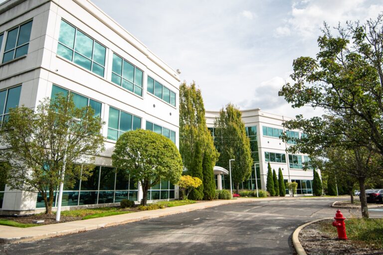 A modern office building with large glass windows surrounded by well-maintained landscaping, including several green trees and shrubs. The building exterior is white, and there is an empty parking lot in the foreground. A red fire hydrant is visible on the right side near the sidewalk.