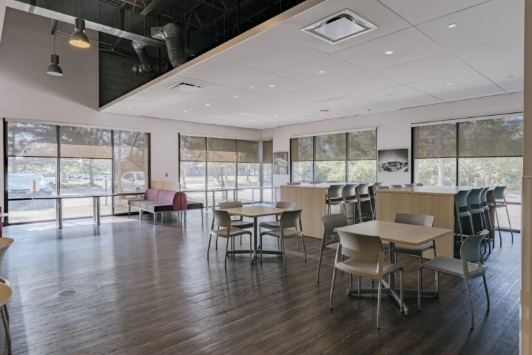A spacious, modern office cafeteria with wooden flooring and large windows. The room features several tables with chairs, a counter with stools, and a small lounge area with sofas. Ceiling lights and exposed ductwork are visible, and natural light filters through the shaded windows, showcasing a view of trees outside.