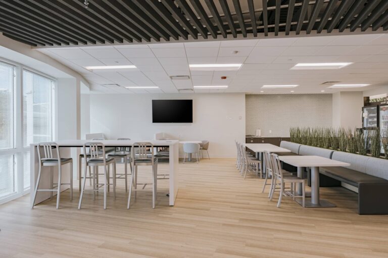 A modern office cafeteria featuring high tables with metal chairs and a seating area with a long bench, small square tables, additional chairs, and decorative plants. The room has a light wood floor, white walls, and a mounted flat-screen television. Ceiling panels and overhead lighting create a bright and open atmosphere.