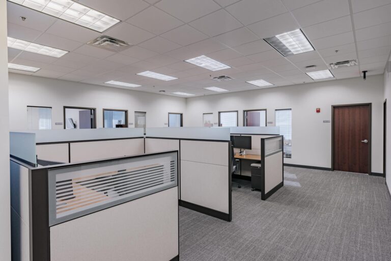 A spacious office interior featuring cubicle workstations with light-colored panels and wooden work surfaces. The room has a neutral color scheme with gray carpet flooring and several closed office doors along the walls. Large overhead fluorescent lights provide bright illumination.