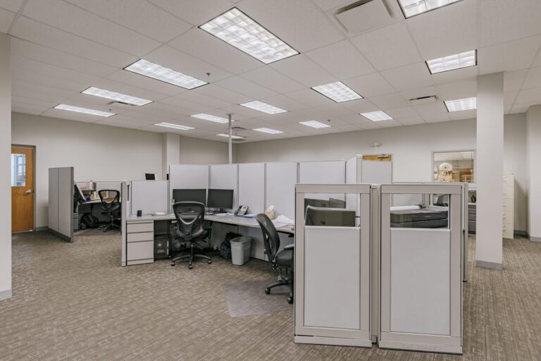 A modern office space featuring several cubicles with white partitions, office chairs, and desks equipped with computer monitors. The room is well-lit with overhead fluorescent lighting and has neutral-toned carpeting. There is a door on the left side and a window on the right showing an additional office area.