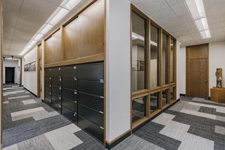 A modern office hallway featuring patterned carpeting, large wooden cabinets, black filing cabinets, and a glass-walled room. The corridor is brightly lit with ceiling lights, and artwork is visible on the walls.