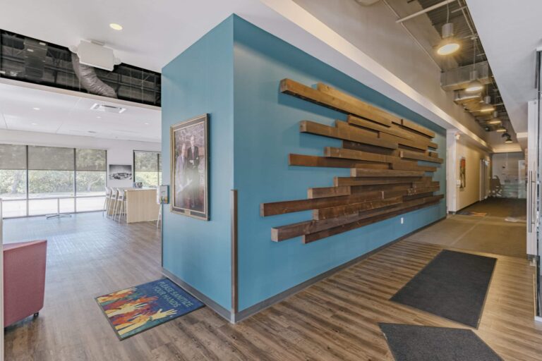 A modern office hallway with a blue wall featuring wooden art installations. The floor is wooden, and there are mats placed near the entrance. A framed picture is hung on the wall, and the space is well-lit with ceiling lights. To the left, there's a seating area with large windows providing natural light.