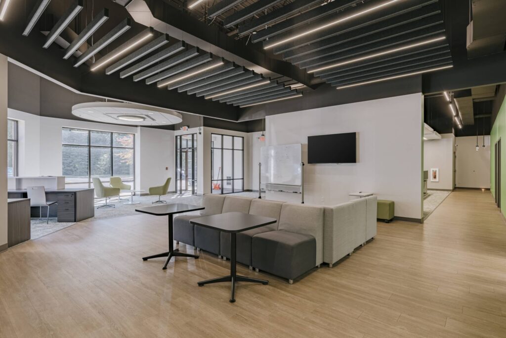 A spacious modern office with an open layout, featuring a light wood floor, dark acoustic ceiling panels, and contemporary lighting. The room includes comfortable grey and green seating areas, a whiteboard, a wall-mounted TV, and several tables. Large windows provide natural light, and glass doors are visible in the background.