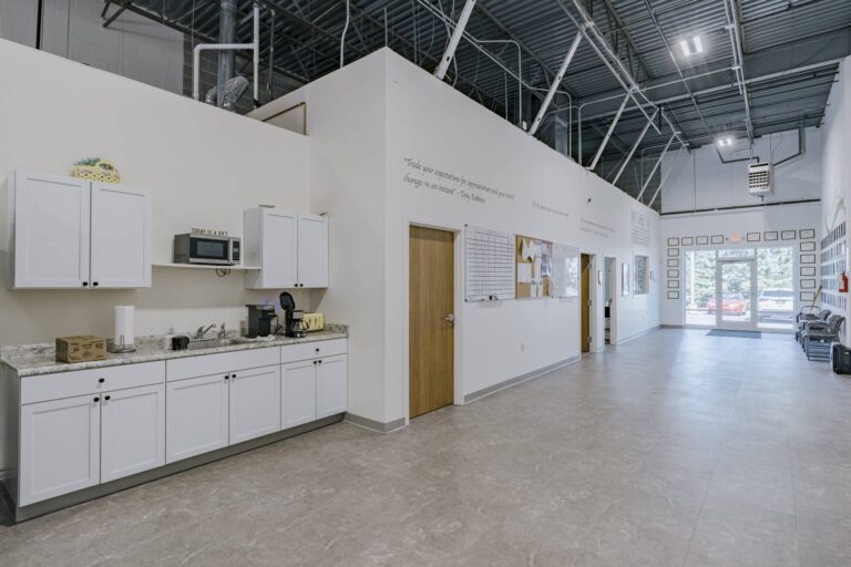 A spacious office kitchen area with white cabinets, a microwave labeled 'Today is a gift,' a coffee maker, and a brown package on the countertop. Adjacent is a hallway with wall decor and several framed items. The area has a high ceiling with exposed beams and a glass door leading outside, flanked by windows and seating on the right.