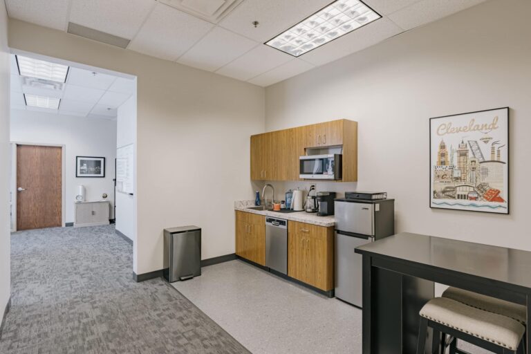 A modern office kitchen area featuring wooden cabinets, a countertop with a sink, a microwave, coffeemaker, and a small refrigerator. In the corner of the room, there is a trash can. A colorful poster labeled 'Cleveland' decorates the wall. Adjacent to the kitchen area is a hallway leading to a doorway and a cabinet with framed artwork above it.