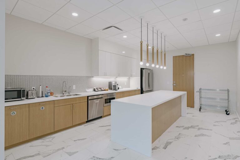 A modern office kitchen with white cabinetry and a wooden island. The kitchen is equipped with stainless steel appliances including a microwave, fridge, and dishwasher. It features white marble floor tiles and pendant lighting above the island. A recycling bin, toaster, coffee maker, and cleaning supplies are visible on the counter.