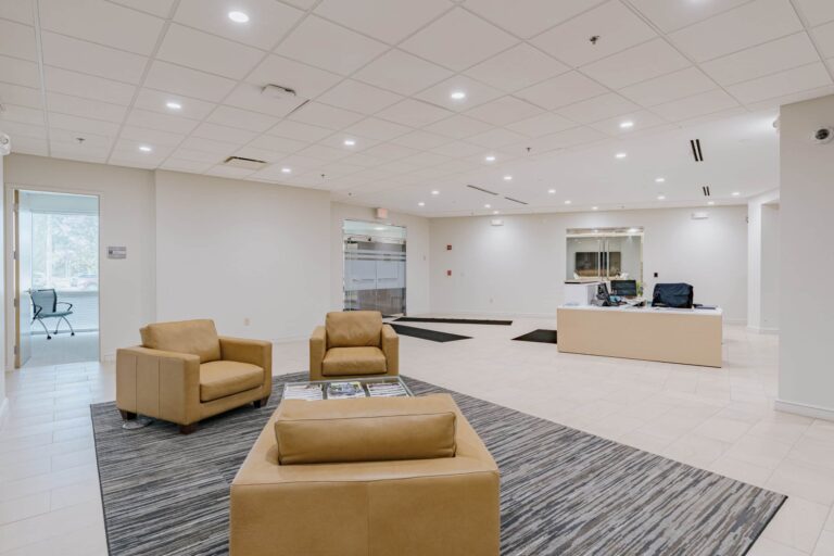 A modern office lobby featuring beige leather chairs around a glass coffee table on a patterned grey rug. The lobby has a tiled floor, a reception desk with office equipment, and glass doors leading to a small meeting room with a chair visible. Bright overhead lighting and a clean, minimalistic design dominate the space.
