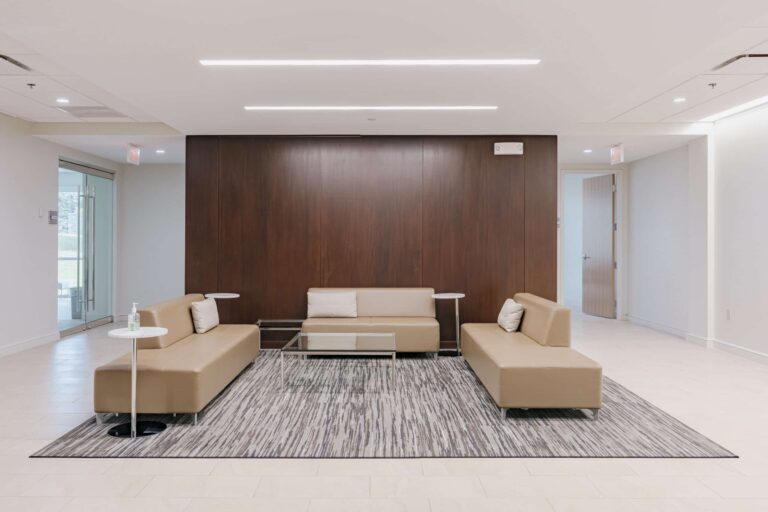 A modern office lobby featuring three beige leather sofas arranged in a U-shape on a textured area rug. The sofas are positioned against a dark wooden accent wall with a rectangular design. Two small white side tables are placed beside the sofas, one with a bottle of hand sanitizer. The floor is light-colored tile, and the ceiling is fitted with recessed lighting. A glass office door is visible on the left, and a wooden door is partially open on the right, showcasing a bright, minimalist space.