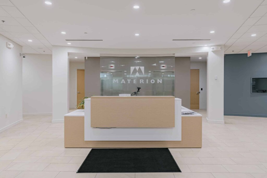 A modern office lobby with a reception desk in the center. The backdrop features the word 'Materion', with glass elements and wooden accents on the desk. The area is well-lit with ceiling lights, and there are doors on either side of the reception area.