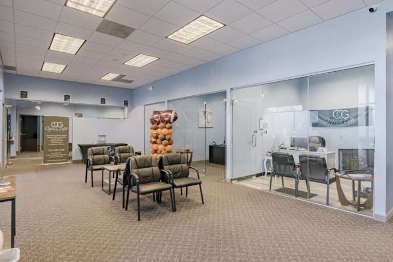 A modern office lobby featuring several black cushioned chairs and a small table arranged in a seating area. The space has carpet flooring and light blue walls. There are glass doors leading to private office spaces. A banner and a standee with the logo of CCG Office Lofts are visible, along with a decorative column of balloons in orange, brown, and black. The ceiling is fitted with recessed lighting panels.