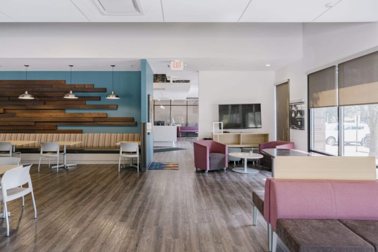 A modern office lounge area featuring wooden flooring and a mix of contemporary furniture. The left side has a wooden accented wall with hanging pendant lights, booths, and white chairs with tables. On the right, there are red and gray cushioned chairs around small tables, and a large flat-screen TV mounted on a light wood cabinet. Large windows provide natural light to the space.
