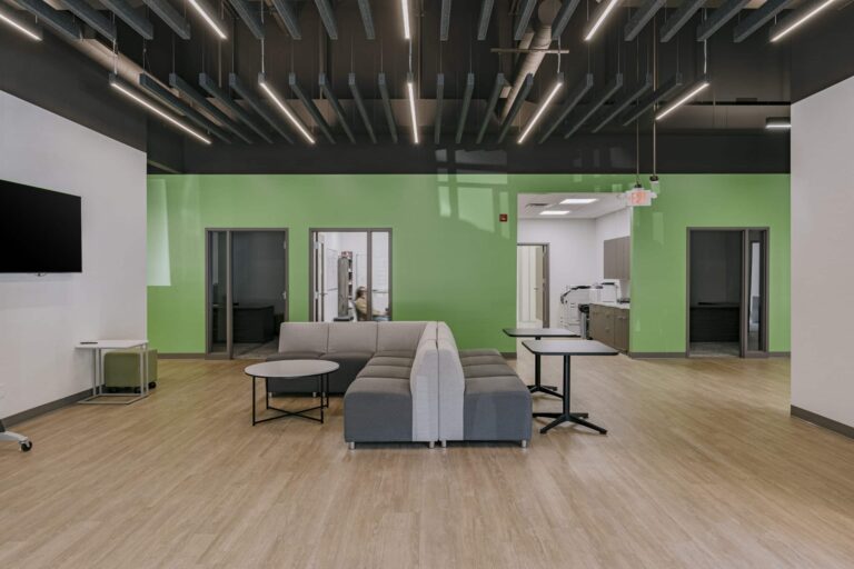 A modern office lounge area featuring a large, gray sectional sofa with attached small round and square tables. The room has wooden flooring, a green accent wall, and a ceiling with linear lights. To the left is a wall-mounted TV and a small table. In the background, there are gray doors and an office copier is visible in a room with white walls.