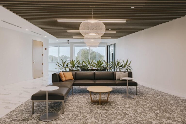 A modern office lounge features a sleek black sectional sofa adorned with orange and gray cushions, flanked by two small round tables. The area is highlighted by pendant lights and a series of potted plants behind the sofa. The ceiling has wooden slats, and large windows provide natural light to the space. The floor is covered with a patterned rug, adding a contemporary touch to the decor.
