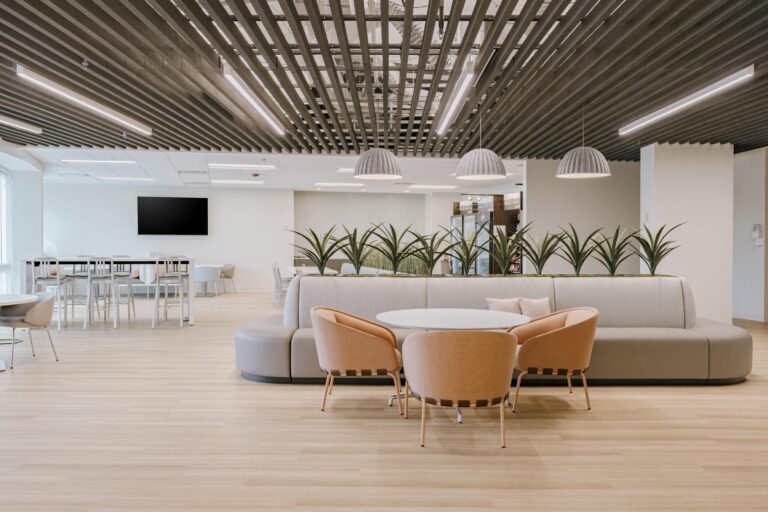 A modern office lounge with a light wood floor and black slatted ceiling. The space features a round table with four orange chairs and a semi-circular gray sofa, adorned with indoor plants behind it. In the background, tall white tables and chairs are visible, along with a large black TV screen mounted on the wall.