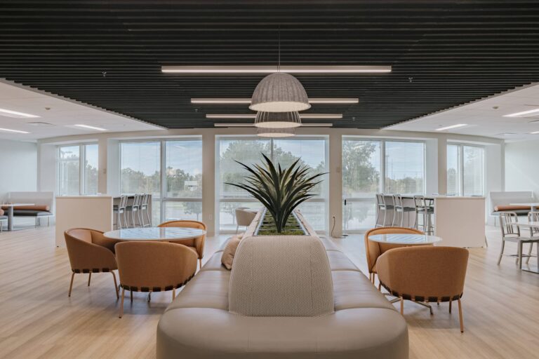 A modern office lounge featuring a central seating area with beige and orange chairs around a round coffee table. A large plant is placed in the middle, creating a natural barrier between seating sections. The room is well-lit with large windows and a high ceiling with dark slats and hanging lights. Additional seating areas and bar stools are visible in the background.