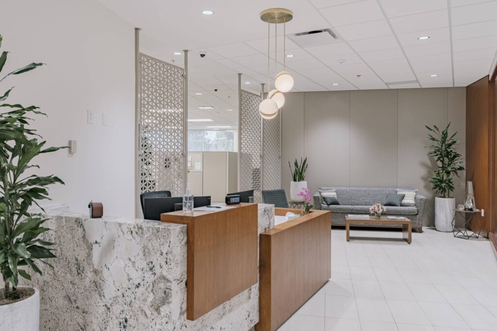 A modern office reception area featuring a granite and wood front desk with two chairs, decorative lighting above, and a seating area with a gray sofa, throw pillows, a wooden coffee table with a flower arrangement, and potted plants on either side.