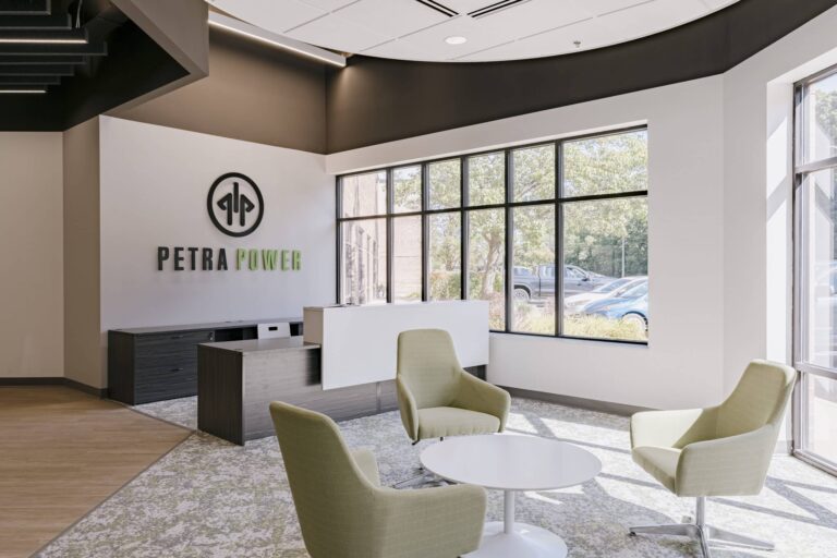 A modern office reception area featuring the Petra Power logo on the wall. The room includes a white desk, three green upholstered chairs, and a round white coffee table. Large windows allow natural light to fill the space, offering a view of trees and parked cars outside. The decor is minimalistic with a combination of wood and carpet flooring.