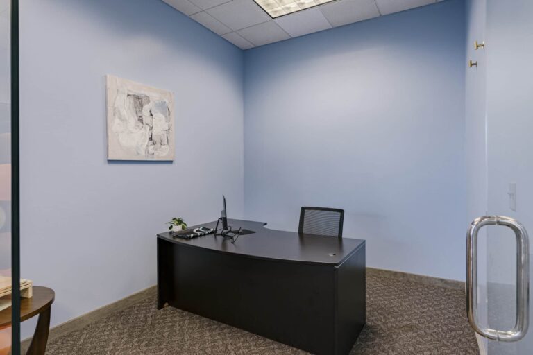 A small modern office room with blue walls, featuring a simple black desk with a monitor, phone, and small potted plant. A black mesh chair is tucked under the desk. On the left wall hangs an abstract artwork. The room has a glass door and a carpeted floor.