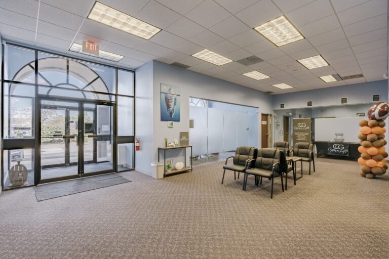 A spacious office waiting area with large glass doors leading outside. Inside, there's a seating arrangement with black chairs and small tables. A decorative table on the left has a small plant and brochures. The walls feature an artwork of an iceberg, and there's a cluster of orange and brown balloons shaped like a football near a reception desk with a sign displaying 'Office Loft'.