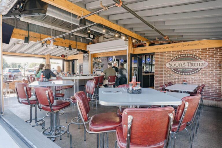The image shows a modern restaurant interior with exposed beams and ductwork. There are high-top tables surrounded by red cushioned chairs, and a bar area with patrons seated. Natural light enters through large open windows, creating a bright atmosphere. The restaurant's name, 'Yours Truly Restaurant,' is visible on a brick wall adjacent to the seating area.