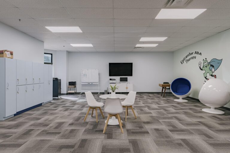A modern therapy office space featuring a mix of contemporary furniture. There is a round table with four white chairs in the center, lockers on the left, and a TV mounted on the wall with a cabinet beneath it. A blue egg chair and a white spherical chair are positioned against a wall with the words 'Epicenter ABA Therapy' and a cartoon character. The room has a neutral color scheme with gray carpet tiles, and a large whiteboard is visible near the TV.