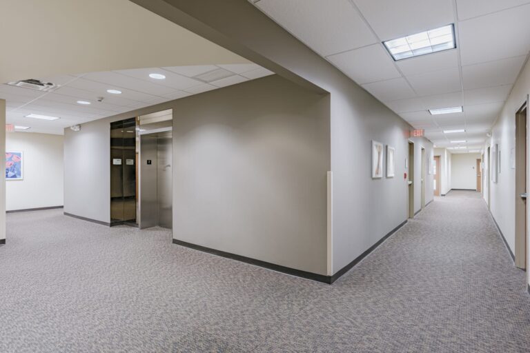 A corridor inside an office building featuring an elevator with metallic doors on the left and a long hallway to the right. The walls are painted white and adorned with framed artwork. The ceiling is fitted with fluorescent lights, and the floor is carpeted. An exit sign is visible at the end of the hallway.