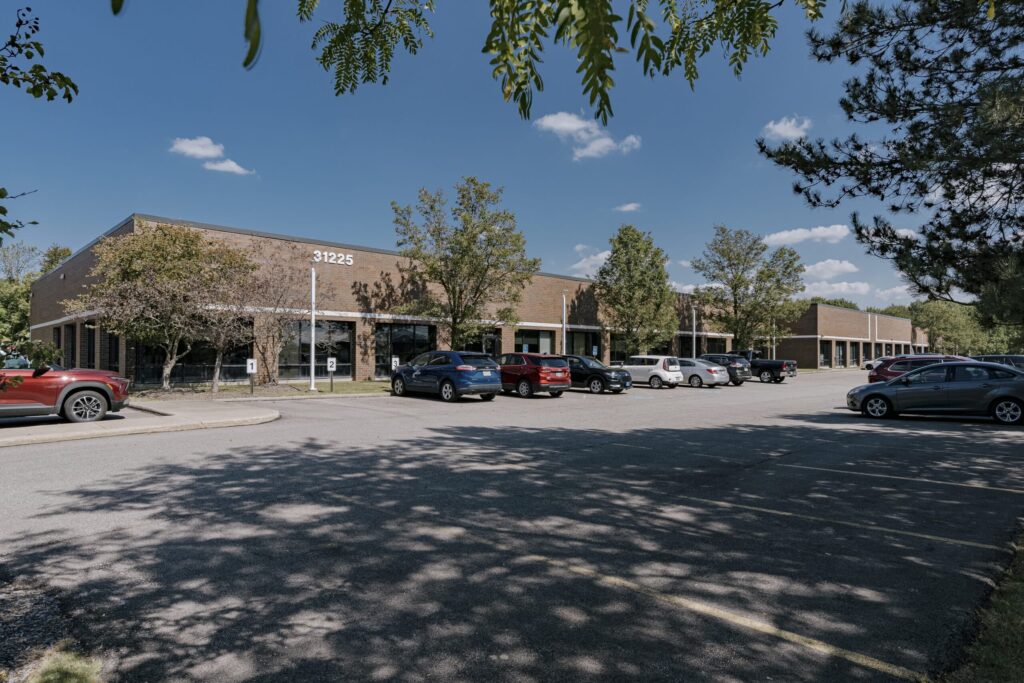 A one-story brick office building with the number 31225 on it, surrounded by a parking lot filled with cars. The area is bordered by trees under a clear blue sky with a few clouds, casting shadows on the pavement.