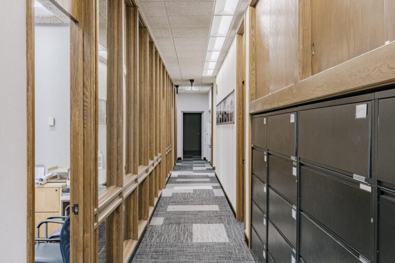 A modern office hallway with gray patterned carpet, wooden framing on the left with a visible office space, and dark filing cabinets lining the right wall. The ceiling features fluorescent lights, and the hallway leads to a dark door at the end.