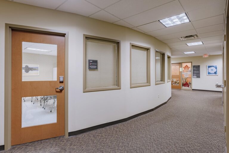 A curved office hallway with multiple closed doors. One door on the left has a window showing chairs and tables inside. There are several windows with blinds drawn and another door at the end with decorations and artwork on the wall.