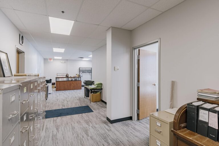 A spacious office hallway with beige walls and ceiling lights. On the left side, there are multiple metal filing cabinets. To the right, an open wooden door leads to another room. At the end of the hallway, a reception desk is visible, alongside a coat rack. The floor is carpeted with a light gray pattern, and a small blue mat is placed on the floor.