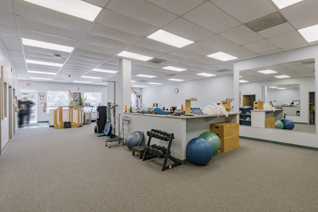 Interior of a physical therapy gym featuring exercise equipment like dumbbells, yoga balls, and physical therapy tools. The room has a carpeted floor, mirrors on the walls, and fluorescent lighting. A person is seen walking towards an exit in the background, and windows allow natural light to enter.