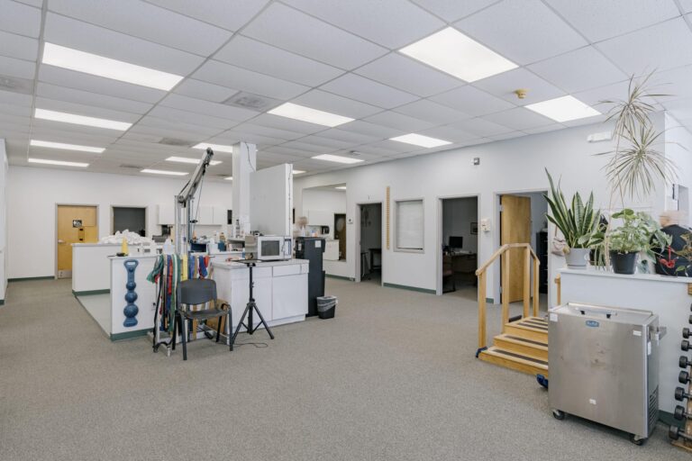 Interior of a physical therapy office featuring open space with exercise equipment, therapy tools, and potted plants. A wooden step platform is visible next to a counter with a microwave, and there are open doorways leading to additional rooms.